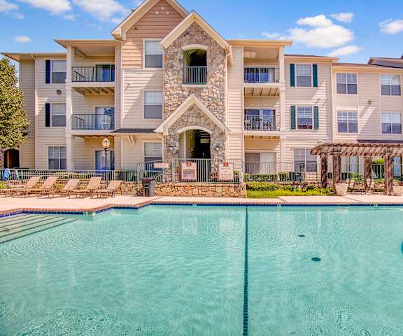 The large, turquoise outdoor pool with pool chairs, tables, and chairs with the apartments in the background at Woodland Park Apartments.