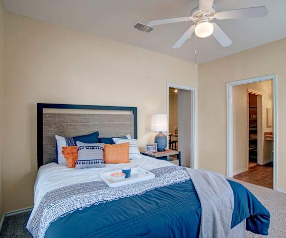 A spacious bedroom with a blue and white queen-size bed with orange accent pillows, overhead lighting, a ceiling fan, decorations, closet, and a nightstand with a lamp at Woodland Park Apartment Homes. 