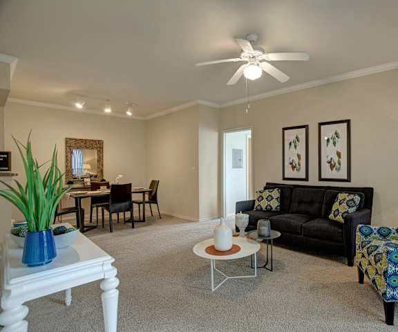 The spacious living room with a couch, plush chairs, coffee table, overhead lighting with a ceiling fan, decorations, a TV, and view of the dining room with a table, chairs, and place settings for four at Woodland Park Apartment Homes. 