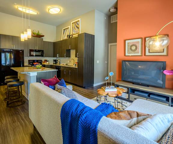 The living room with a couch, plush chairs, coffee table, decorations, a TV, and view of the kitchen at Harmony Luxury Apartments. 