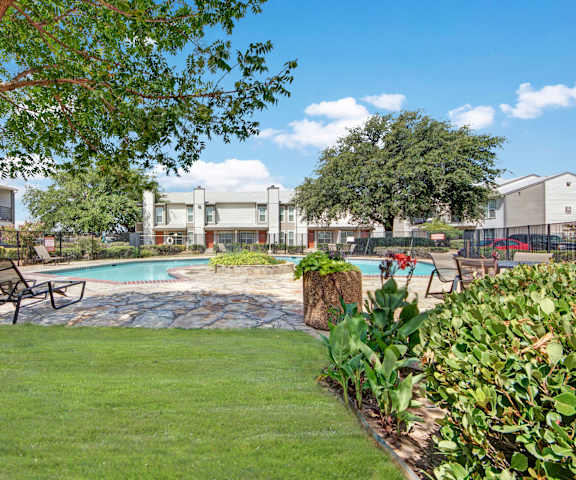 The turquoise outdoor pool with pool chairs, tables, and chairs with the apartments in the background and a grassy area in the foreground at High Plains Apartment Homes. 