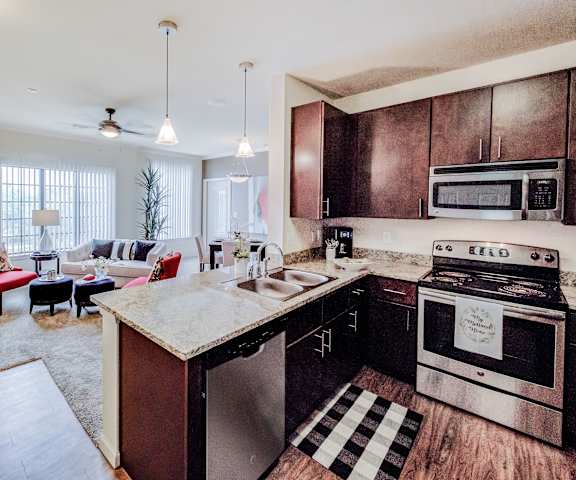 The living room with a couch, plush chairs, coffee table, decorations, a TV, windows, and view of the dining room and the kitchen with stainless steel appliances, dark wood cabinets, and marble counters at Le Mirage Apartments. 