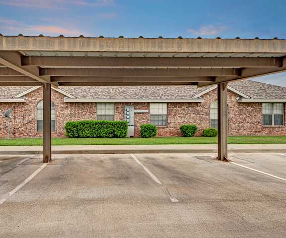 The exterior of the buildings and covered parking lot with grass and bushes in the background at Lexington Court Apartment Homes. 