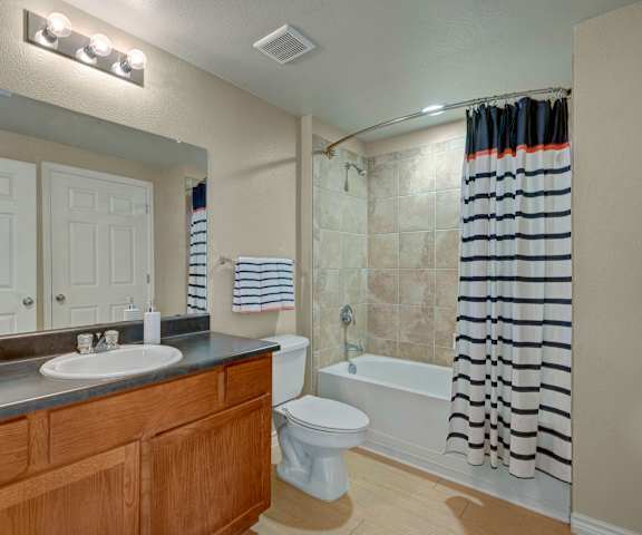 The bathroom with a sink, mirror, lights, toilet, shower, and striped shower curtain at Mission Green Apartments. 