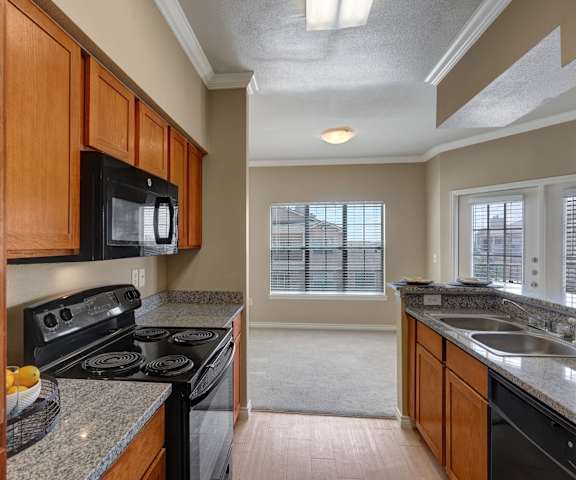 A view of the empty carpeted living room with a window and the kitchen with black appliances and large wooden cabinets at Mission Green Apartments. 