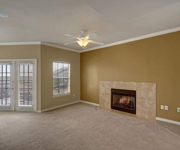 An empty living room with carpeting, a fireplace, an overhead light and ceiling fan, and French doors to the balcony at Mission Green Apartments. 