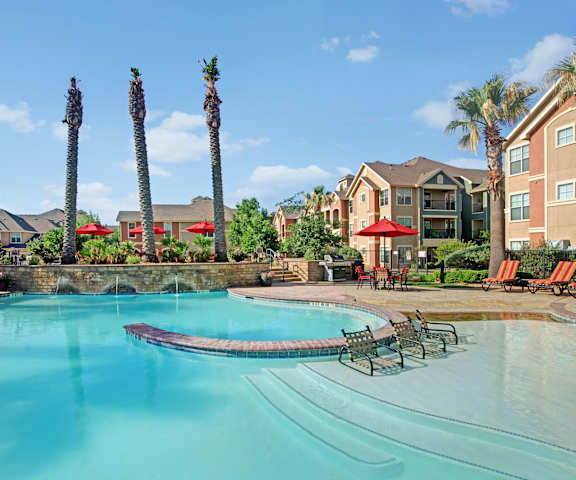 The large, turquoise, resort-style outdoor pool with orange pool chairs and red umbrellas with greenery, palm trees, and the apartments in the background at the Palms at Briarwood Apartment Homes. 