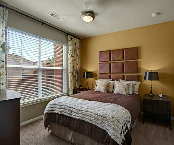A spacious bedroom with a queen-size bed with a brown and white bedspread, a dresser, wall art, lamps, decorations, a large window with curtains, and nightstands with lamps at the Palms at Briarwood Apartment Homes. 