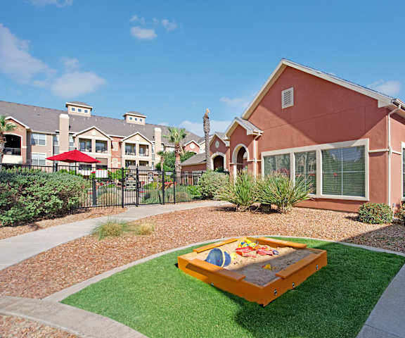 A sandbox for children with toys surrounded by grass and pathways by the leasing office at the Palms at Briarwood Apartment Homes. 