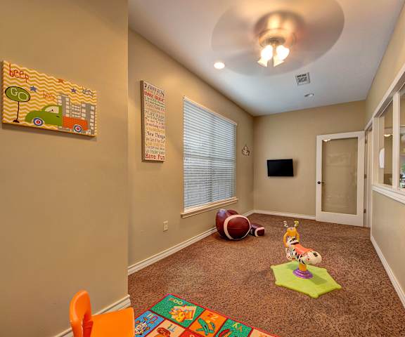 A carpeted playroom with wall art, large windows, overhead lighting and ceiling fan, a TV, and toys on the floor at the Reserve at Abilene Apartment Homes. 