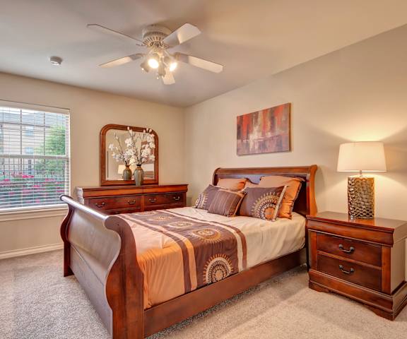 A spacious bedroom with a queen-size bed with a white and brown bedspread, a dresser, wall art, lamps, decorations, a window, overhead lighting with a ceiling fan, and a nightstand with a lamp at the Reserve at Abilene Apartment Homes. 