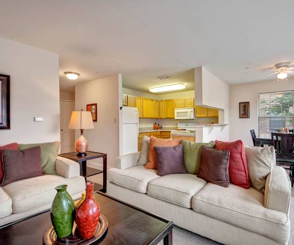 The spacious living room with white couches with colorful pillows, a coffee table, overhead lighting with a ceiling fan, decorations, a lamp on a coffee table, a TV, and view of the dining room with a table, chairs, and the kitchen at the Reserve at Abilene Apartment Homes. 