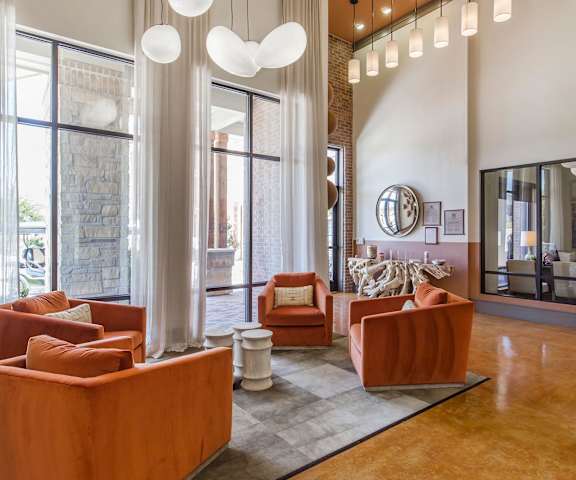The amazing resident lounge with decorations, large windows with white curtains, unique overhead lighting, a TV, orange plush chairs, coffee tables, and a view of the leasing office at Sorrel Fairview Apartment Homes.