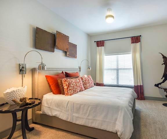 A spacious bedroom with a king-size bed with an orange and white bedspread, a dresser, wall art, lamps, decorations, a large window with blinds and orange and white curtains, and nightstands at Sorrel Fairview Apartment Homes. 