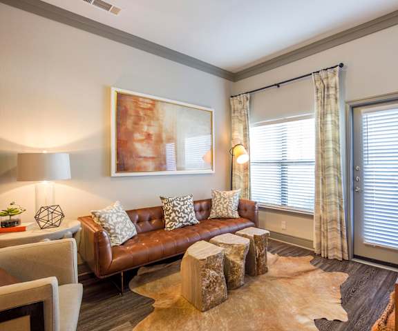 The spacious living room with a brown leather couch, cow printed throw pillows, large windows, pottery, wall art, a white plush chair, coffee table, decorations, large windows, a cowhide rug, and glass doors to the balcony at Sorrel Fairview Apartment Homes. 