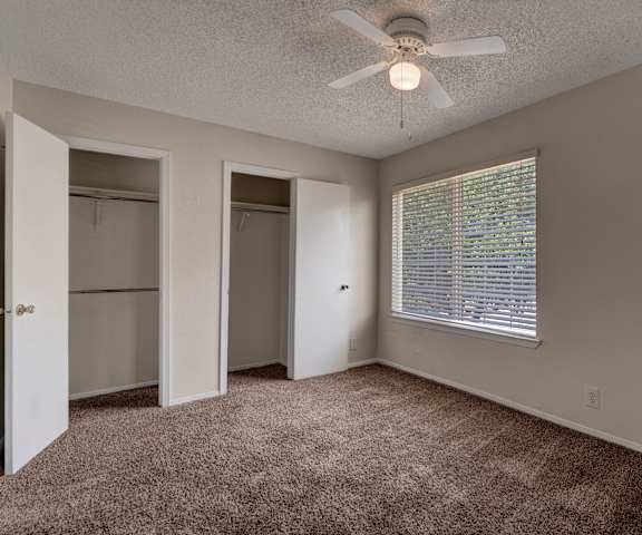 A large, empty carpeted bedroom with overhead lighting, a ceiling fan, a window, and two closets at the Bradford Apartment Homes. 