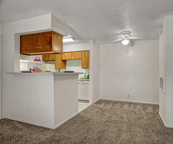 A view of the empty carpeted living room with overhead lighting and a ceiling fan and the kitchen with white appliances and wooden large cabinets at the Bradford Apartment Homes. 