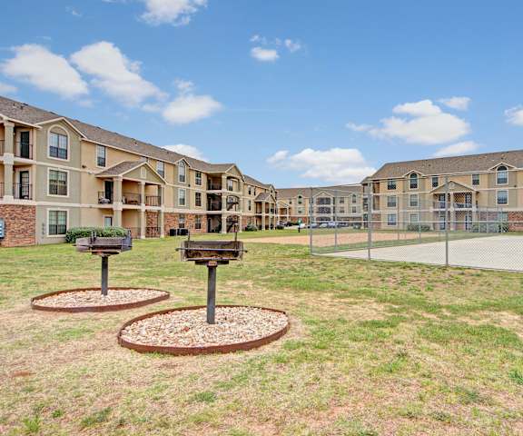 Two community grilling stations with grassy area and basketball court next to them and the tan apartment buildings in the background at the Residence at Heritage Park Apartment Homes. 
