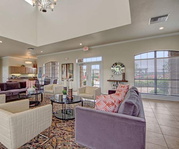 The amazing resident lounge with light purple couches, decorations, large windows, glass doors to the pool, chandeliers, white plush chairs, and coffee tables with a resident kitchen in the background at the Residence at Heritage Park Apartment Homes. 