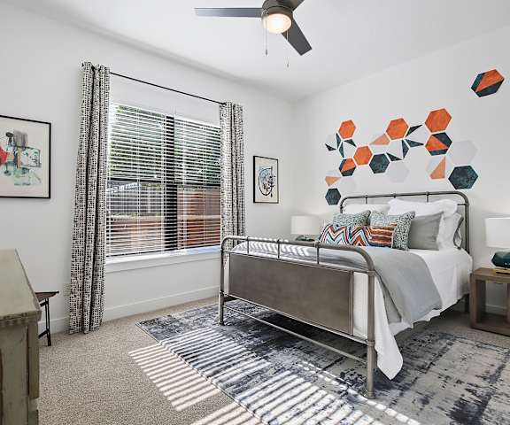 A spacious, gray bedroom with a queen-size bed with a gray and white bedspread, a dresser, wall art, lamps, decorations, window, white, orange, and black hexagon wall art, and nightstands at the Uptown Apartment Homes. 