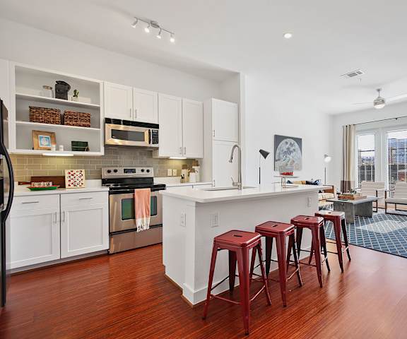 A large, modern kitchen with stainless steel appliances, bar counter with red barstools, kitchen utensils, white marble countertops with lots of room, and large, white wooden cabinets at the Uptown Apartment Homes. 