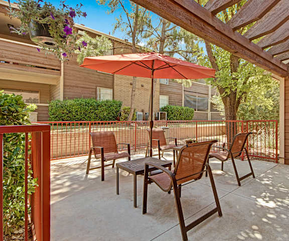 A patio with four chairs, two tables, and an orange umbrella with the apartment buildings and plants in the background. 