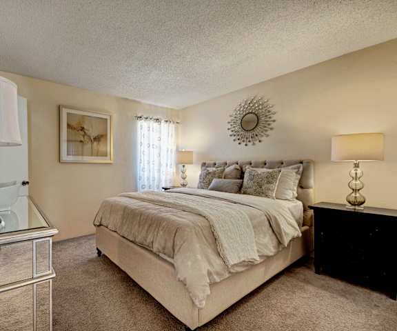 A spacious, ivory bedroom with a queen-size bed with an ivory bedspread, a dresser, wall art, lamps, decorations, window, and nightstands at Trinity Place Apartment Homes. 