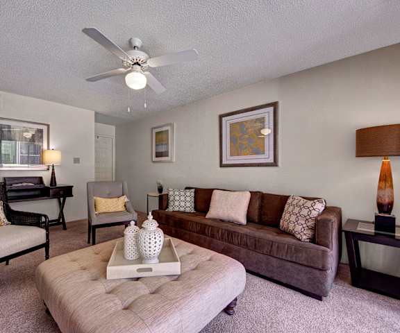 The spacious living room with a brown plush couch, plush white and gray chairs, a white fabric coffee table with a tray on top, overhead lighting with a ceiling fan, wall art, coffee tables, and decorations, at Trinity Place Apartment Homes. 