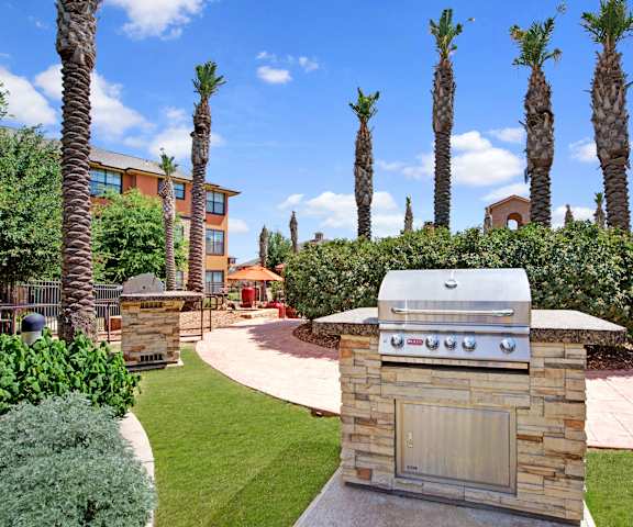 The grilling area surrounded by grass with a grill, bushes, and palm trees in the background at Tuscany at Faudree Apartment Homes. 
