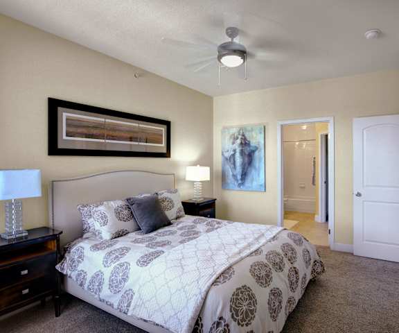 A spacious bedroom with a queen-size bed with a white and brown floral bedspread, a dresser, wall art, decorations, closet, and nightstands at Tuscany at Faudree Apartment Homes. 