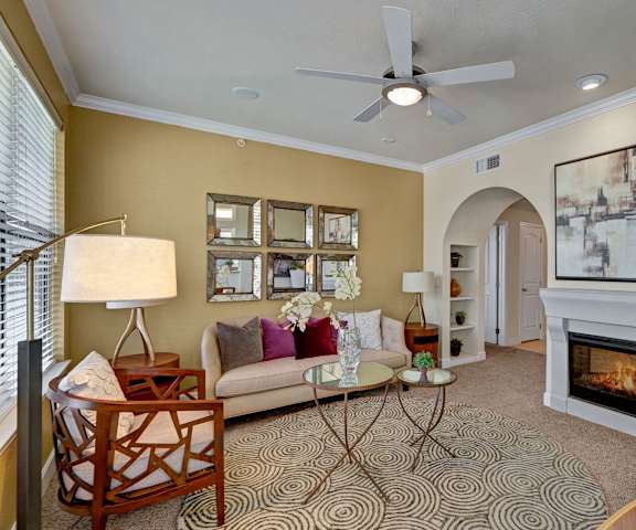 The amazing resident lounge with a fireplace, couches, decorations, large windows with curtains, mirrors on the wall, a ceiling fan, plush chairs, and coffee tables at Tuscany at Faudree Apartment Homes. 