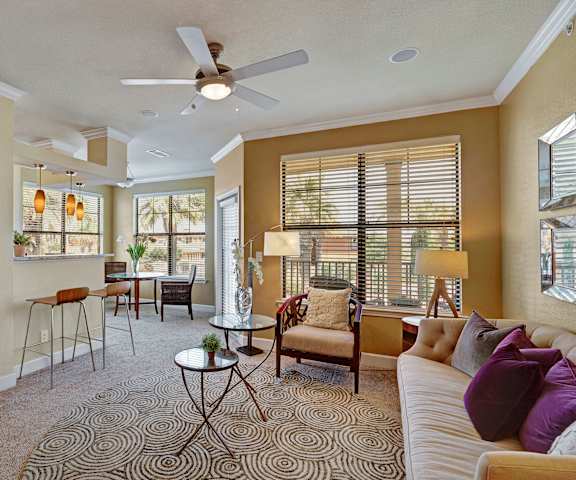 The yellow, spacious living room with a tan couch, plush chairs, coffee table, large windows, overhead lighting with a ceiling fan, decorations, a TV, and view of the dining room with a bar with two barstools at Tuscany at Faudree Apartment Homes. 