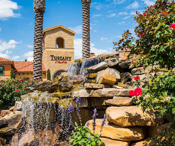 The exterior sign with trees and surrounded by greenery with flowers in front of a cascading waterfall with the buildings in the background of Tuscany at Faudree Apartment Homes. 