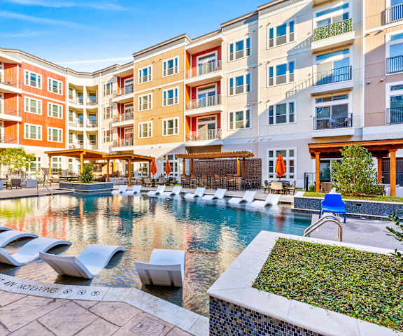 The large, turquoise outdoor pool surrounded by palm trees, pool chairs, tables, cabanas, and chairs with the white, yellow, and orange apartments in the background at Waterford Market Apartment Homes. 