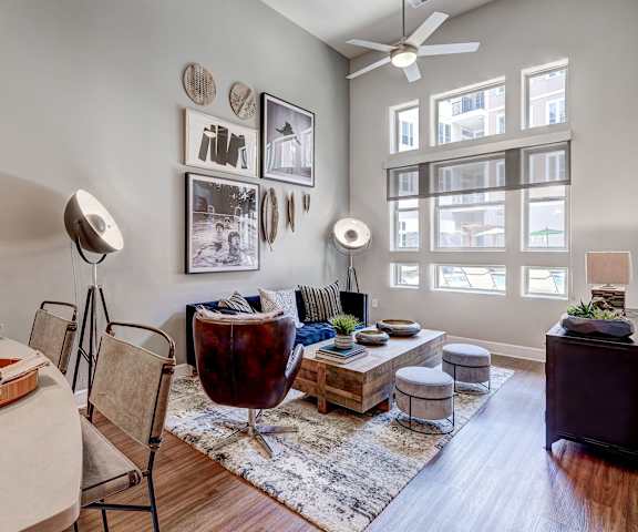 The spacious living room with a couch, throw pillows, large windows, a ceiling fan, wall art, plush chairs, coffee table, decorations, a TV, and glass doors to the balcony at Waterford Market Apartment Homes. 