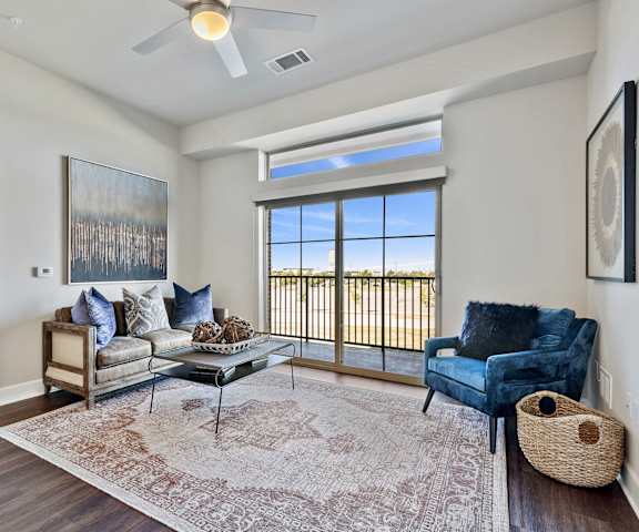 The spacious living room with a couch, throw pillows, large windows, a ceiling fan, wall art, plush chairs, coffee table, decorations, a TV, and glass doors to the balcony at Waterford Market Apartment Homes. 