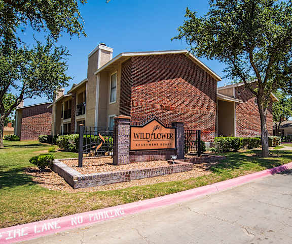 The exterior sign with trees and surrounded by greenery with the red brick buildings in the background of Wildflower Apartment Homes. 