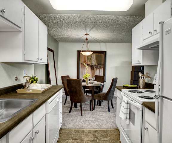 The cozy kitchen with spacious white cabinets, counters with lots of room, and white appliances, and the dining room with plush browns chairs, dining tables, and large mirrors on the wall at Overlook on Magnolia Apartments. 