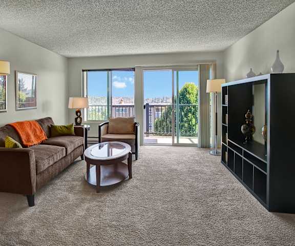 The spacious living room with a couch with bright pillows and an orange blanket, a plush chair, coffee table, decorations, an entertainment system, and balcony at Overlook on Magnolia Apartments. 