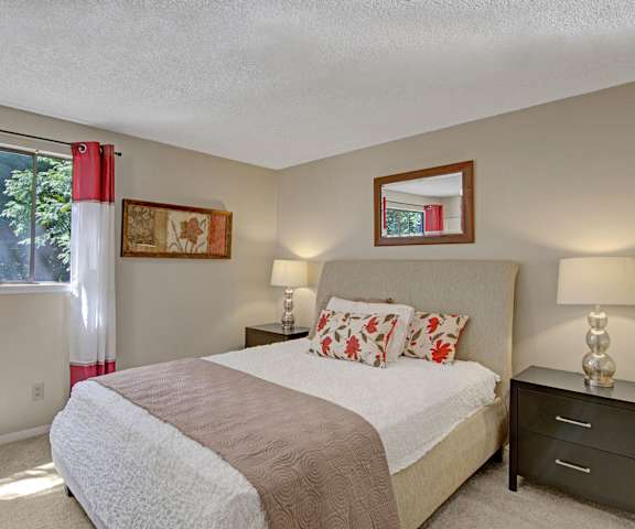 A spacious bedroom with a queen-size bed with a tan and white bedspread, a mirror, wall art, lamps, decorations, window with curtains, and nightstands at the Ridge and Shores Apartment Homes. 