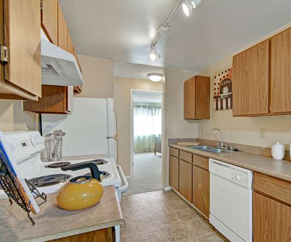 A cozy kitchen with white appliances, wall art, kitchen utensils, overhead lighting, light countertops with lots of room, and large wooden cabinets at the Ridge and Shores Apartment Homes. 