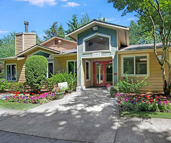 The exterior of the leasing office showing a blue and yellow house with flower beds, trees, and grass in front at the Ridge and Shores Apartment Homes. 