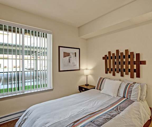 A spacious bedroom with a queen-size bed with a white striped bedspread, a dresser, wall art, lamps, decorations, window, and nightstands with lamps at Riverside 9 Apartment Homes. 
