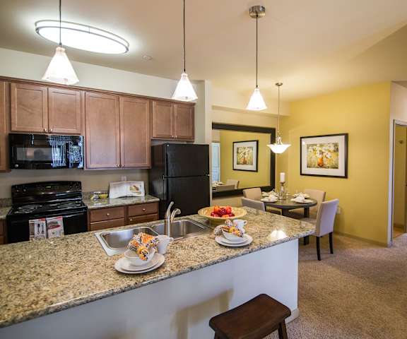 A spacious kitchen with black appliances, marble countertops with lots of room, and wooden cabinets with decorations and a view of the yellow dining room with a table, four chairs, and four places set at Riverside 9 Apartment Homes. 