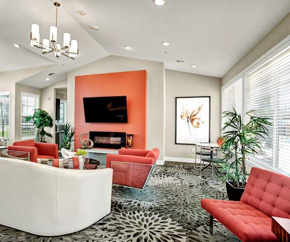 The amazing resident lounge with a fireplace, TV, white and red couches, decorations, large windows with curtains, orange accent wall, decorative plants, orange plush chairs, and coffee tables at Solara Apartment Homes. 