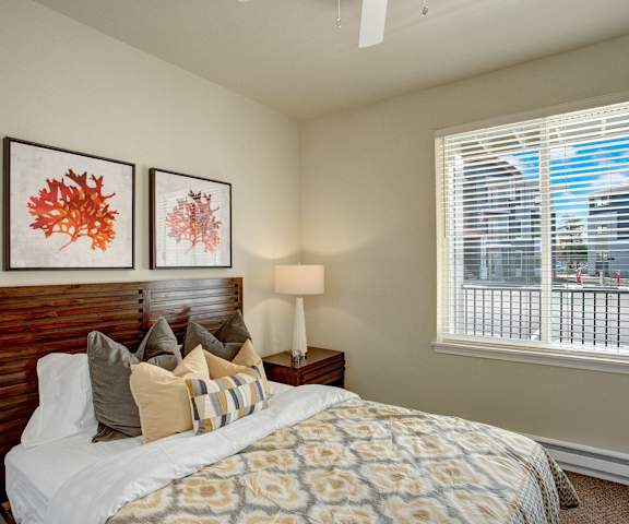 A spacious bedroom with a queen-size bed with a white, gray, and yellow bedspread, a dresser, wall art, lamp, decorations, window, and nightstand at Solara Apartment Homes. 
