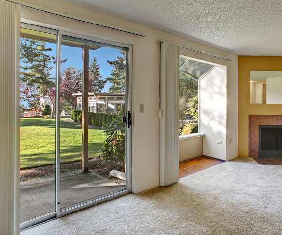 A large, empty living room with carpeting, a fireplace with a mirror above it, and a door to the patio with the grassy area and trees in the background at Soundview Apartment Homes. 