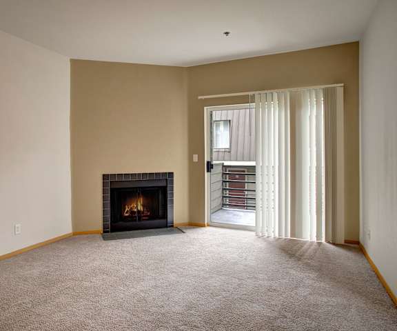 A large, empty living room with carpeting, a fireplace, and a sliding glass door with white curtains to the balcony at St. James Tower Apartment Homes. 