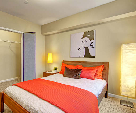 A spacious bedroom with a queen-size bed with a white and orange bedspread, a dresser, wall art, decorations, closet, and nightstands at the Artiste Apartment Homes. 