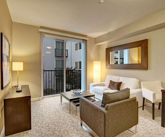 The spacious living room with a white couch, brown throw pillows, a mirror, wall art, plush chair, coffee table, decorations, a TV, and glass doors to the balcony at the Artiste Apartment Homes. 